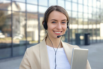 Portrait of smiling young caucasian employee in headphones with microphone look at camera standing near the businesscentre after work. Freelance business women.