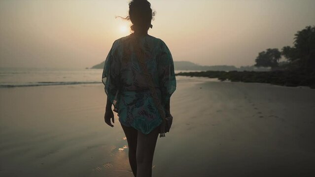 Landscape view of a young woman walking on a sandy tropical beach by the ocean, towards the sunset