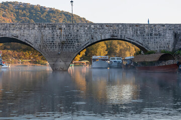Beautiful view of medieval stone arch bridge over Crmnica river in Lake Skadar National Park in idyllic village Virpazar, Montenegro, Balkans, Europe. Small boats floating on the calm water surface