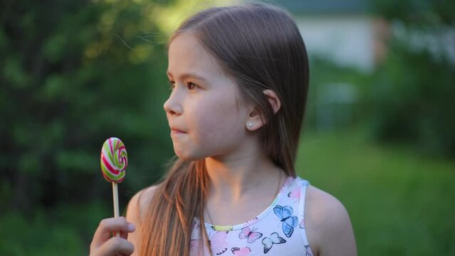 Close-up front view of pretty brunette Caucasian girl with brown eyes looking at camera licking multicolored lollipop in slow motion. Happy satisfied child posing in summer park enjoying tasty dessert