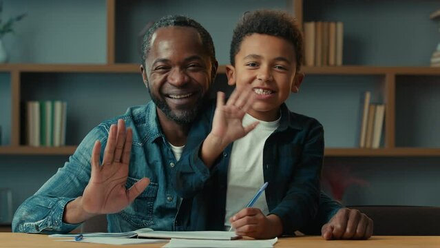 African American father dad with little son kid child boy schoolboy schoolchild schoolkid sit at table after homework writing home learning education looking to camera waving hello greeting smiling