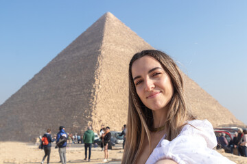 Young happy smiling woman taking a selfie in the pyramids of Cairo, Egypt. With tourist people in the blurred background - obrazy, fototapety, plakaty