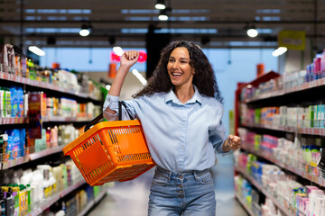 Portrait of a happy woman shopper in a supermarket, a Hispanic woman with a basket of goods smiles with pleasure and dances among the shelves with goods, a satisfied shop customer.