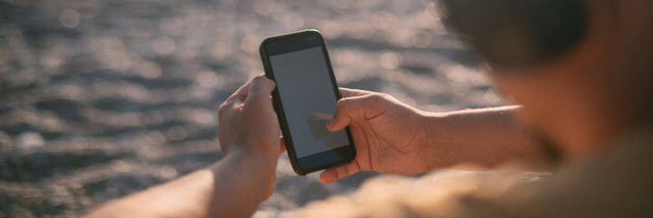 Portrait of a young man sitting on the seashore wearing headphones with a phone in his hands at sunset. © Anna