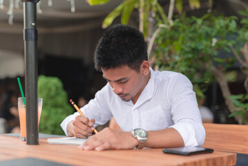 A male illustrator draws an artwork using a pencil and a blank sketchpad while dining at an outdoor...