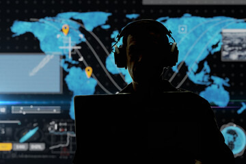 Fototapeta Silhouette of a military man in headphones at a laptop against the background of a world map, contour lighting. Concept: collection of confidential information, surveillance and control of people. obraz
