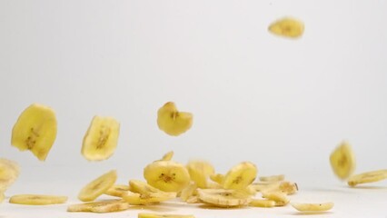 Crispy yellow freeze dried banana chips falling down, spinning and bouncing into a pile on white table top in slow motion