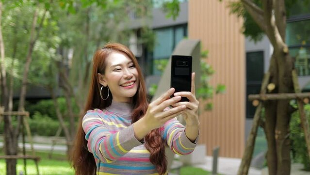 Cheerful Asian woman happy She walks with her smartphone taking photos and video calls online in the outdoor garden of her modern apartment. Housing concept. city ​​living