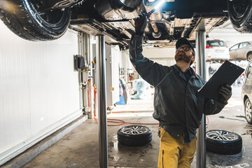 Mechanic with a clipboard inspecting the car chassis of a lifted car using a torch. High-quality photo