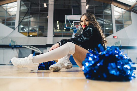 Horizontal full shot of a cheerleader in black and white uniform sitting on basketball court. Blue shiny pom-pom blurred in the foreground. High quality photo