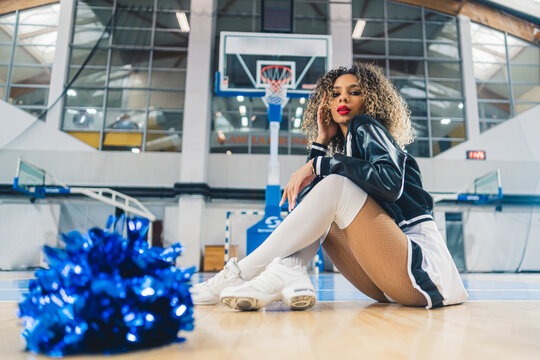 Horizontal shot of cheerleader with curly hair in black jumper sitting on basketball court. Blue shiny pom-pom blurred in the foreground. High quality photo