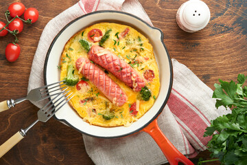 Omelette or frittata with two sausages, melted cheese, cherry tomatoes, broccoli and parsley on red skillet or iron pan on old wooden rustic table background. Healthy breakfast food concept. Top view