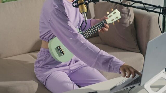 Asian gen Z girl playing ukulele, singing in mic and using laptop with midi controller while recording track at home music studio