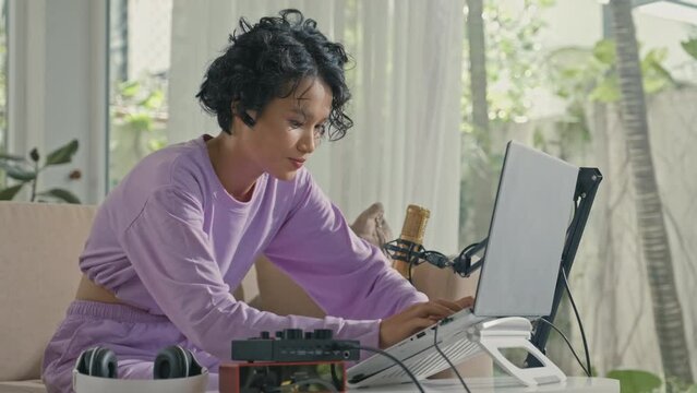 Medium shot of Asian gen Z girl typing on laptop while creating audio content at home music studio equipped with recording setup