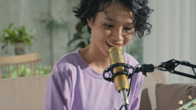 Tilt up shot of Asian gen Z girl playing ukulele and singing in mic while recording song at home music studio