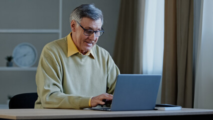 Old Caucasian man with computer work indoors typing chatting writing book online text. Elderly mature senior businessman worker entrepreneur grandfather at office at home at table with laptop working