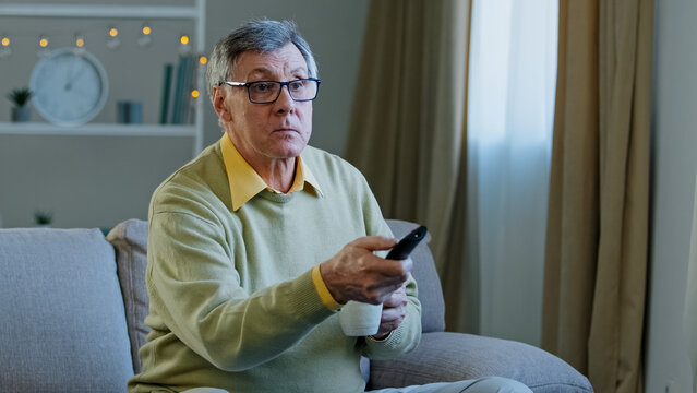 Caucasian Aged Senior Mature Man In Glasses Relax On Couch At Home Change Channel With Remote Controller Watching TV Program Relaxing With Drinking Tea Coffee Old Male Choose Movie Television Indoors