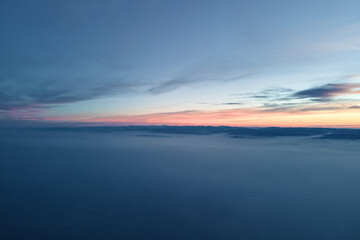 Aerial view of colorful sunset over white dense foggy clouds cover with distant dark silhouettes of mountain hills on horizon