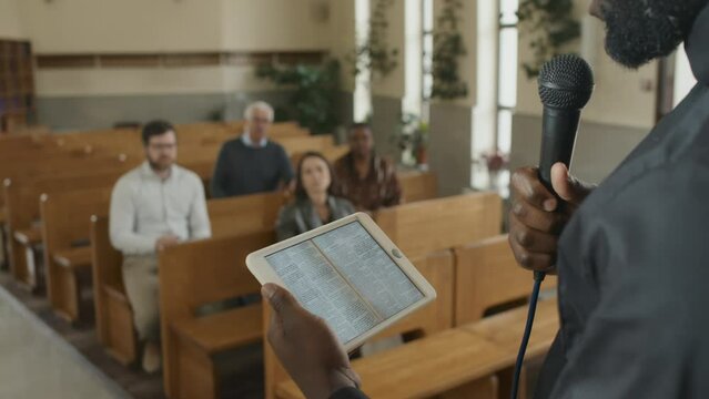 Selective focus shot of modern Catholic pastor holding digital tablet with the Bible book on screen preaching to parishioners