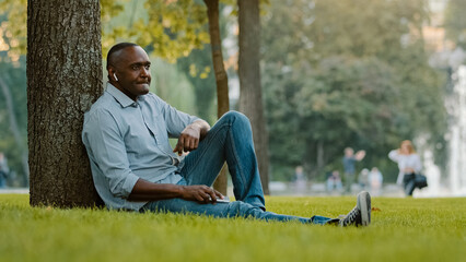 Calm mature adult male enjoying weekend outdoors listening song in wireless headphones with great sound. Senior middle-aged African American man sitting on grass in park listen music with mobile phone