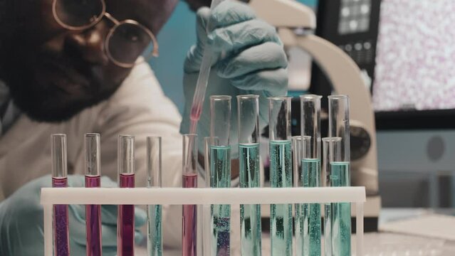 Timelapse close-up of young African American male scientist in gloves and lab coat carrying out scientific research or experiment while working with liquids in flasks by his workplace