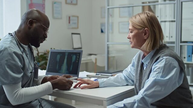 Mature female patient sitting in front of African American general practitioner putting pulse oximeter on her fingertip during check-up of oxygen level in her blood