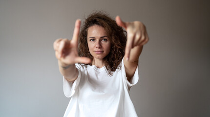 Young brunette curly hair woman wearing white tee shirt looking at camera through her fingers against plain grey background.