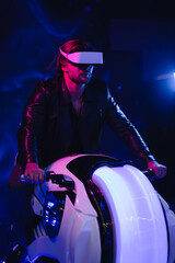 A handsome man in virtual reality glasses, in the dark sits on a space motorcycle. A guy in a leather jacket in the scenery of the future in neon color flies through space.