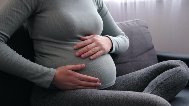 Close up of young pregnant woman sitting on comfortable sofa, puts palms on big belly feel unconditional love to unborn baby. Third trimester of pregnancy, happy maternity, expectant mother concept