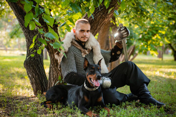 A young man in chain mail with a sword in the forest sits under a tree with a dog. Warrior in armor with a doberman on a chain.