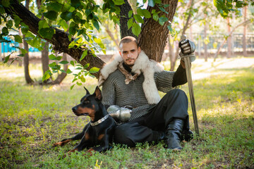 A young man in chain mail with a sword in the forest sits under a tree with a dog. Warrior in armor...