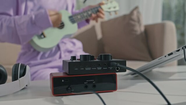 Rack focus tilt up shot of Asian gen Z girl using midi controller and laptop and then playing ukulele and singing in mic while creating music at home recording studio