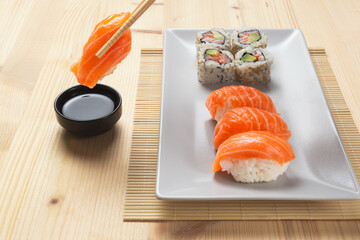 Chopsticks dipping a nigiri salmon piece on a black bowl with soy sauce and a white plate with various sushi over a wooden table