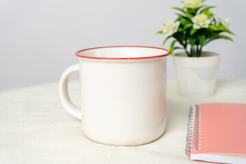 A white blank enamel mug on the top of a white table with some books decorated behind it, minimalistic looks