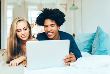 .Young beautiful interracial couple relaxed lying down in bed on the computer