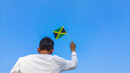 Boy holding Jamaica flag against clear blue sky. Man hand waving Jamaican flag view from back, copy...