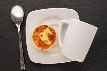 One homemade cheese pancake on white ceramic saucer with white cup and metal spoon on slate stone, macro, top view.