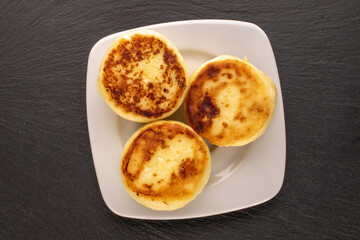 Three homemade cheese pancakes on a white ceramic saucer on slate stone, macro, top view.