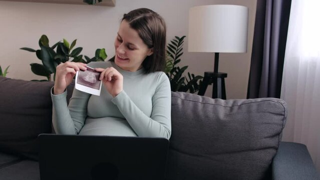 Happy beautiful young pregnant woman showing photo of baby during online call with family by pc laptop while sitting on cozy sofa. Smiling future mother talking via video link from pc computer at home