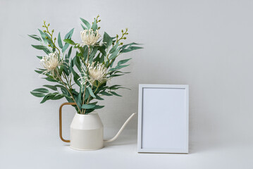 Elegant minimalistic composition with flower bouquet in watering can and empty white photo frame on light neutral background, spring aesthetic template with copy space