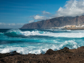 Big breaking waves splash against black lava rock coast. Turquoise blue Atlantic ocean with view of Los Gigantes cliffs and village. Tenerife, Canary islands, Spain. Sunny winter day.
