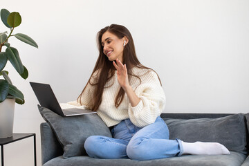 Fototapeta A young woman is sitting on a sofa, smiling and waving during an online meeting using a laptop.  obraz