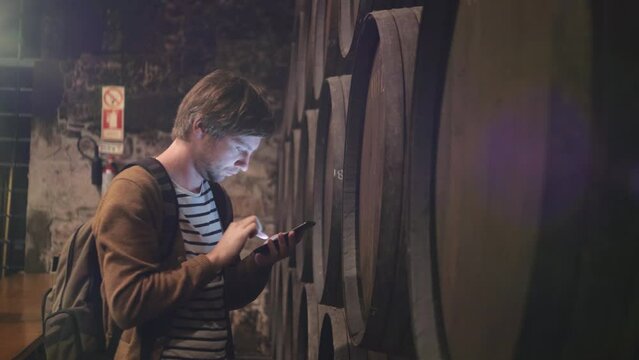 Man winemaker at winery checking barrels in wine cellar with phone and tablet pc.
