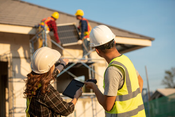 Young technician standing on metal platform installing heavy solar photo voltaic panel on blue sky...