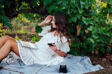 a beautiful woman sits under a grape with a laptop and a glass of red wine