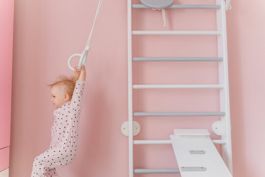 A  toddler girl in soft pink pajamas does gymnastic at home wall bars