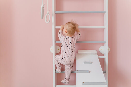 A  toddler girl in soft pink pajamas does gymnastic at home wall bars