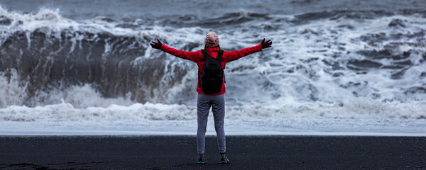 Fearless strong woman with outstretched arms faces powerful waves on black beach; concept of strong...