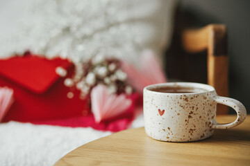 Obraz na płótnie Canvas Cute stylish cup of tea with heart on wooden table against modern armchair with gifts, red envelope and white flowers. Valentine morning surprise for beloved. Happy valentines day!