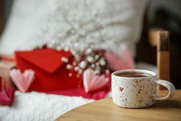 Cute stylish cup of tea with heart on wooden table against modern armchair with gifts, red envelope...
